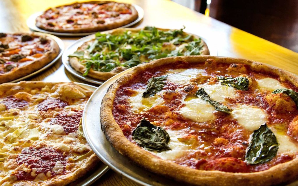 An image of a table with 4 pizzas on it, all baked with melted slightly browned cheese and assorted toppings, and they all look great!