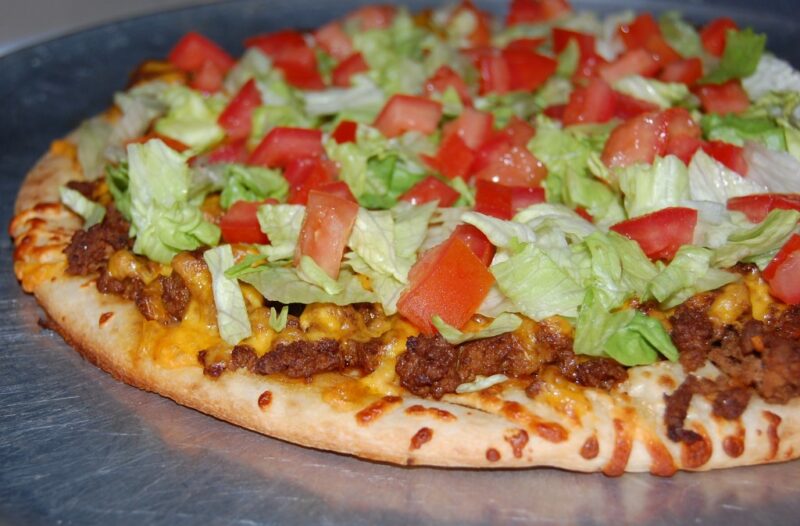 An Image of baked Large Taco Pizza topped with Seasoned Ground Beef, Cheddar Cheese, Roma Tomatoes, Ice Berg Lettuce, and a drizzle of Sour Cream.