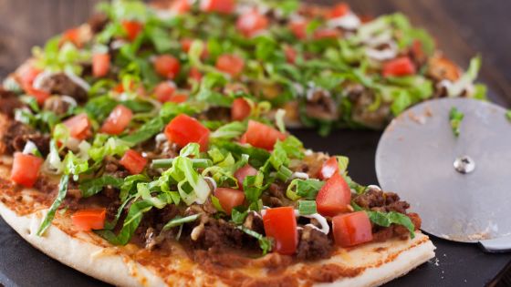 An Image of beautifully baked Large Taco Pizza topped with Meat, Cheese, Tomatoes, Lettuce, and a drizzle of sour cream.